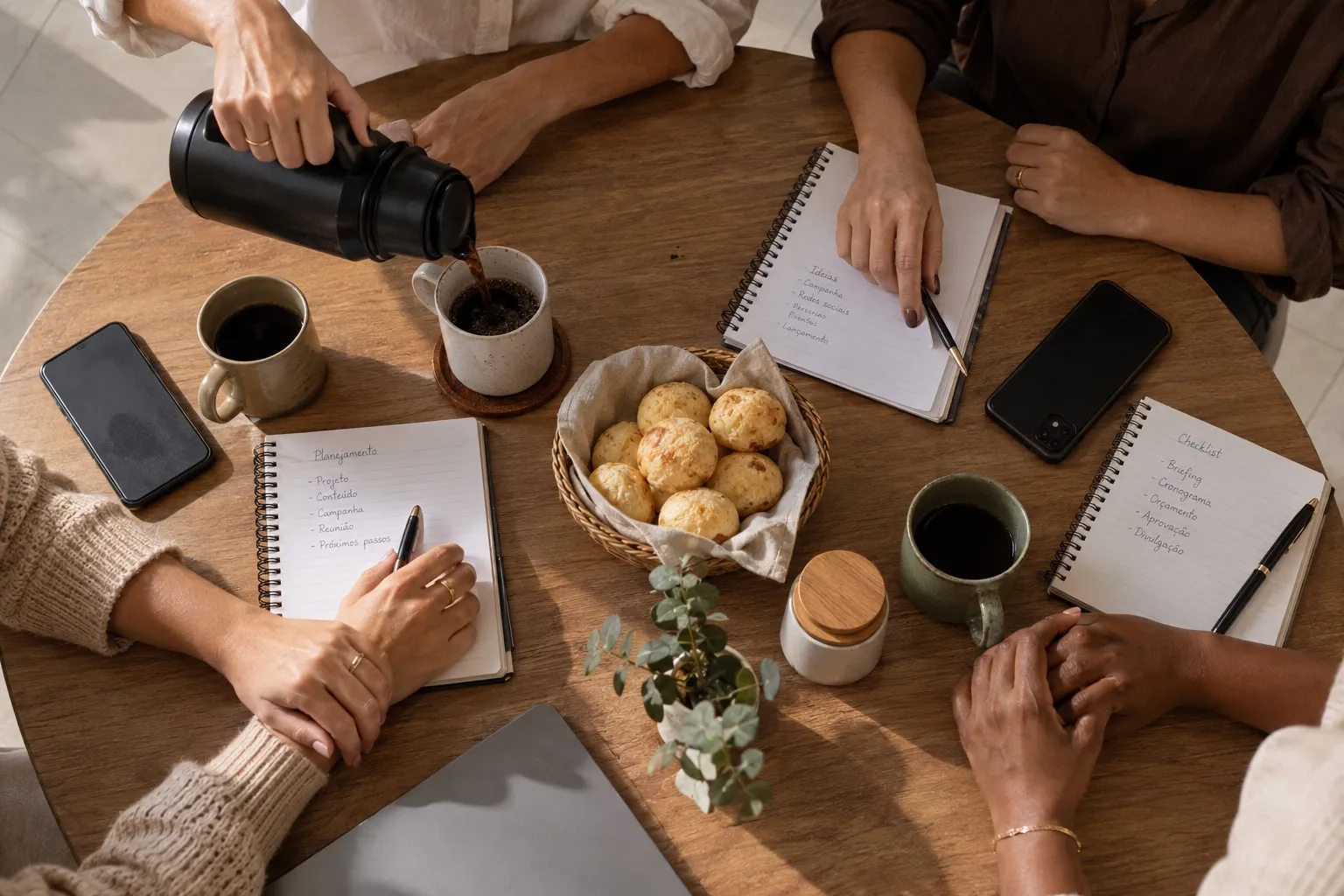 Mãos de psicólogas em volta de uma mesa com cafés, cadernos e pão de queijo